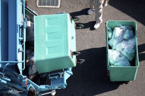 An overhead view of a waste collection scene showing a partially visible blue waste collection vehicle with an open rear compaction area on the left side of the image, parked on a grey asphalt surface. Next to the vehicle, there is a large, green, wheeled waste bin with a textured lid, positioned in the centre of the image, filled with mixed rubbish. To the right, another green bin, slightly smaller, is filled with tightly packed plastic bags containing waste, some of which are transparent, revealing various types of discarded materials. Part of a person's white trousers and shoes are visible in the top right corner, standing on the pavement near the waste bins. The background shows minimal surroundings, focusing mainly on the waste collection and disposal process, suggesting an outdoor area in Camden or nearby residential or commercial premises, as handled by Office Clearance Camden.