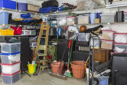 A cluttered storage area or garage filled with various plastic storage boxes, some transparent and others opaque, stacked along shelves and on the floor. The plastic boxes have different colours, including blue, red, white, and black, and contain items that are not visible. There are several cleaning tools such as a broom, mop, and a yellow bucket positioned in the foreground, leaning against the stacked containers. A small wooden ladder is placed at an angle against the shelves, with a few wooden chairs stacked nearby. The environment appears well-lit, with a concrete floor and metal shelving units holding additional clutter, including a sports ball and miscellaneous household items. The scene likely depicts a space in Camden used for storing household or office clearance rubbish, with visual cues suggesting the need for rubbish removal or clearance services, as offered by Office Clearance Camden.