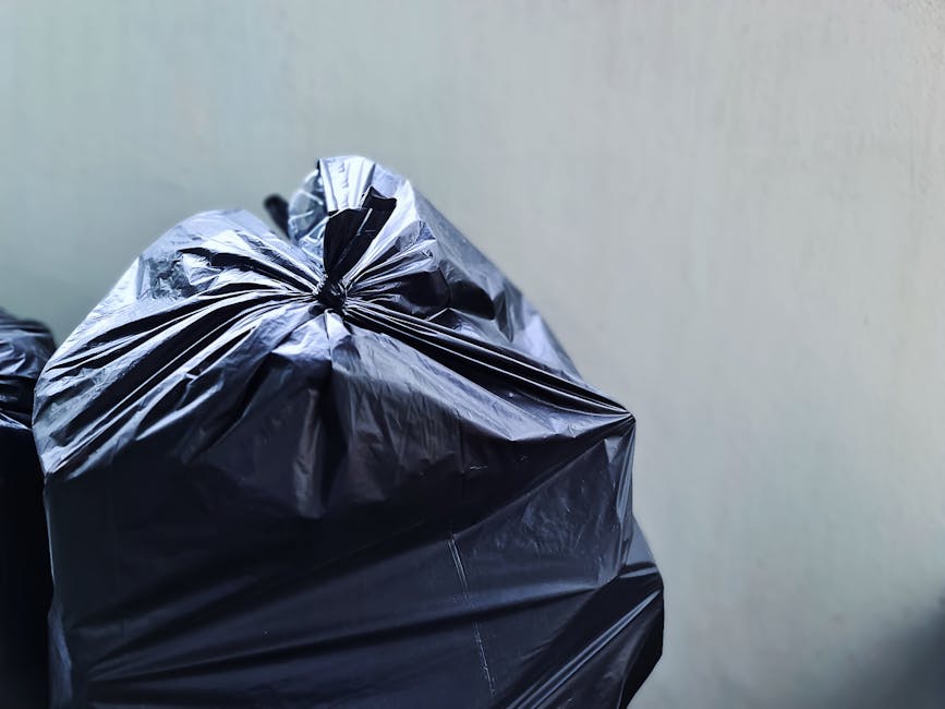 A small, blue utility truck parked on the side of a street during daylight hours, heavily loaded with discarded household items and waste materials in the truck bed, including flattened cardboard boxe
